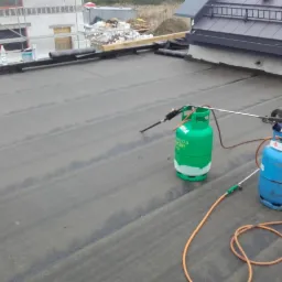 Prace dekarskie: papa termozgrzewalna na płaskim dachu, butle gazowe z palnikami dekarskimi w trakcie łączenia warstw papy, widoczny kubeł z masą bitumiczną i rolka papy. W tle budynek w trakcie...