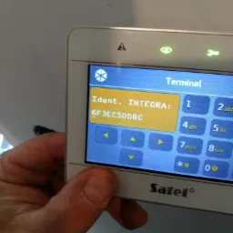 Ekran centrali alarmowej Satel Integra z widocznym identyfikatorem, trzymany w dłoni, widoczne diody sygnalizacyjne i klawiatura numeryczna.