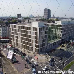 Widok z góry na nowoczesny budynek biurowy z białymi elewacjami i regularnymi oknami, otoczony ulicami z ruchem samochodowym i placem budowy z maszynami, w tle panorama miasta