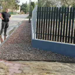 Nowo postawione, czarne, metalowe ogrodzenie z podmurówką w kolorze szarym, obsypane drobnym kamieniem, z pracownikiem w tle, stojącym na mokrym chodniku, widok na ulicę i domy w oddali.