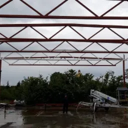 Konstrukcja stalowa z kratownicami w trakcie budowy, widoczna podnośnik koszowy i osoba w oddali na mokrej posadzce.