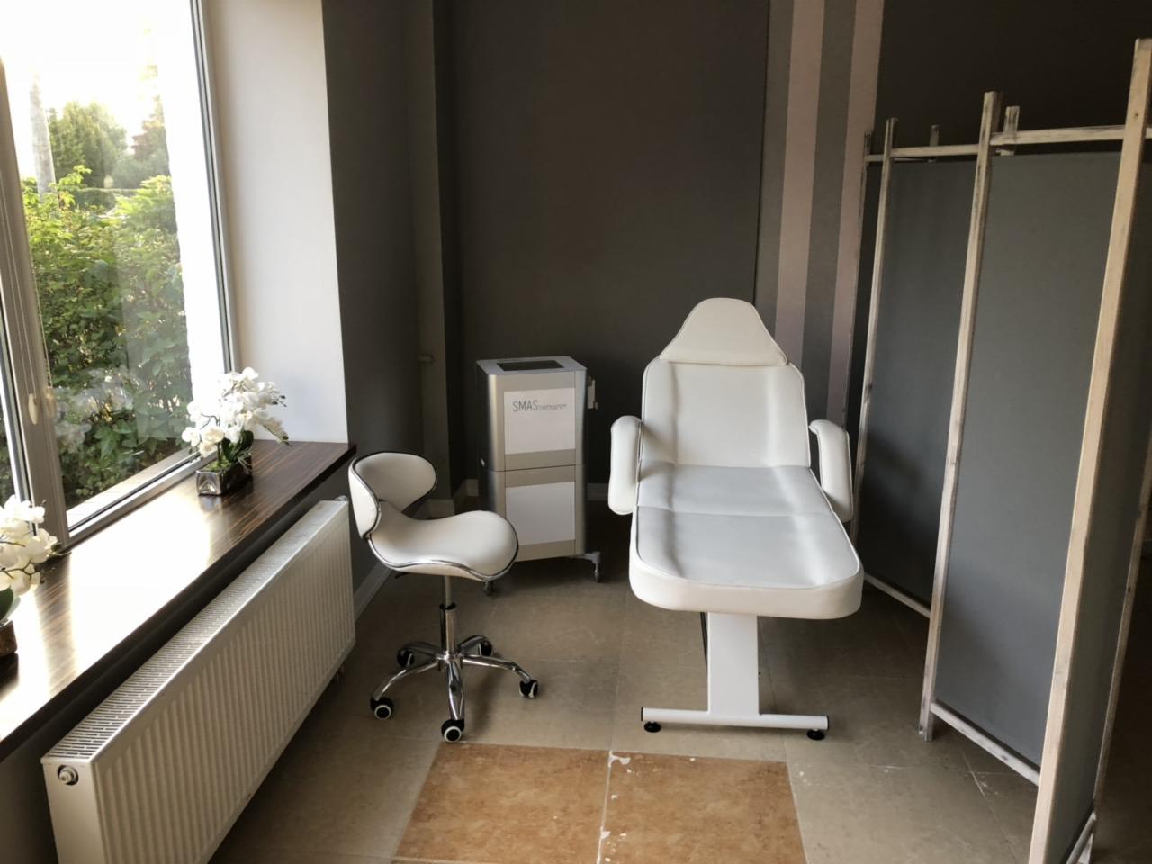 Jasne wnętrze gabinetu z białym fotelem zabiegowym, krzesłem na kółkach, parawanem i sprzętem SMAS w tle, widok z okna z roślinnością.