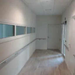 Jasny korytarz biurowy z drewnopodobną podłogą, białymi ścianami, oknami w górnej części ściany i metalowymi poręczami.