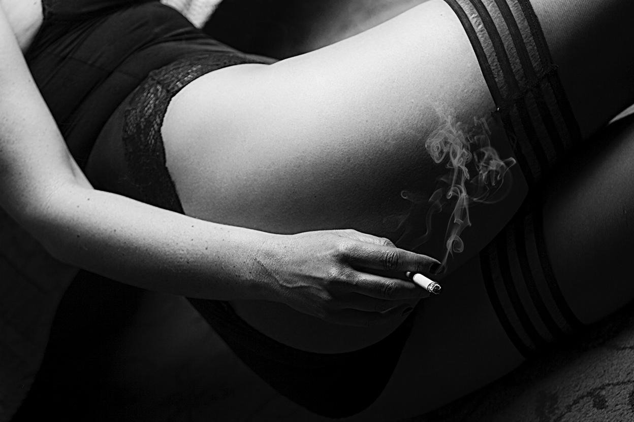 Czarno-białe ujęcie kobiety w bieliźnie i pończochach, leżącej z papierosem w dłoni, z dymem unoszącym się w powietrzu.