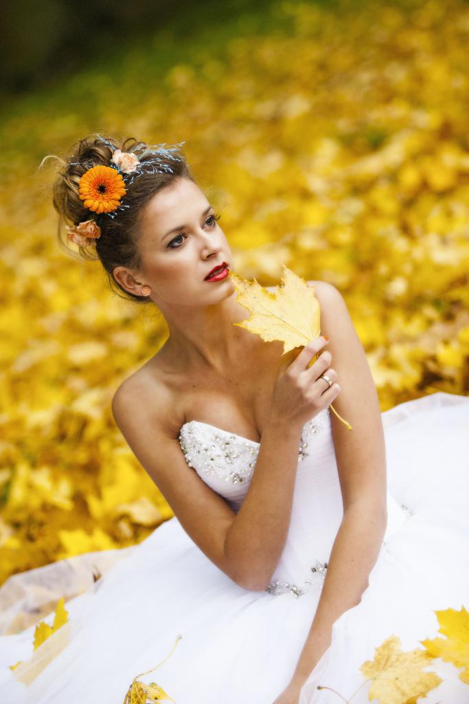 Panna młoda w białej sukni z kryształkami, trzymająca żółty liść na tle opadłych liści, z pomarańczowym kwiatem we włosach i mocną czerwoną szminką.