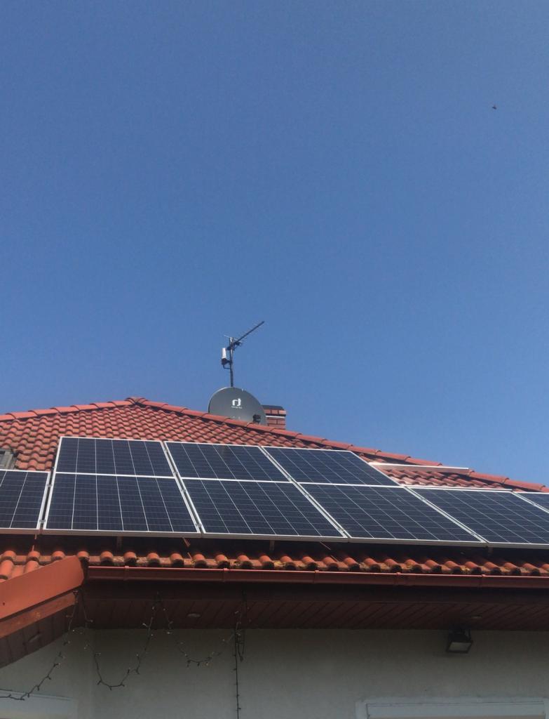 Czerwony dach z panelami słonecznymi w układzie poziomym, antena satelitarna i antena telewizyjna na tle błękitnego nieba.