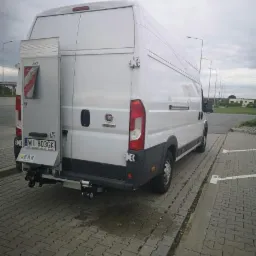 Biały samochód dostawczy z platformą załadunkową z tyłu, zaparkowany na brukowanym parkingu w pochmurny dzień.