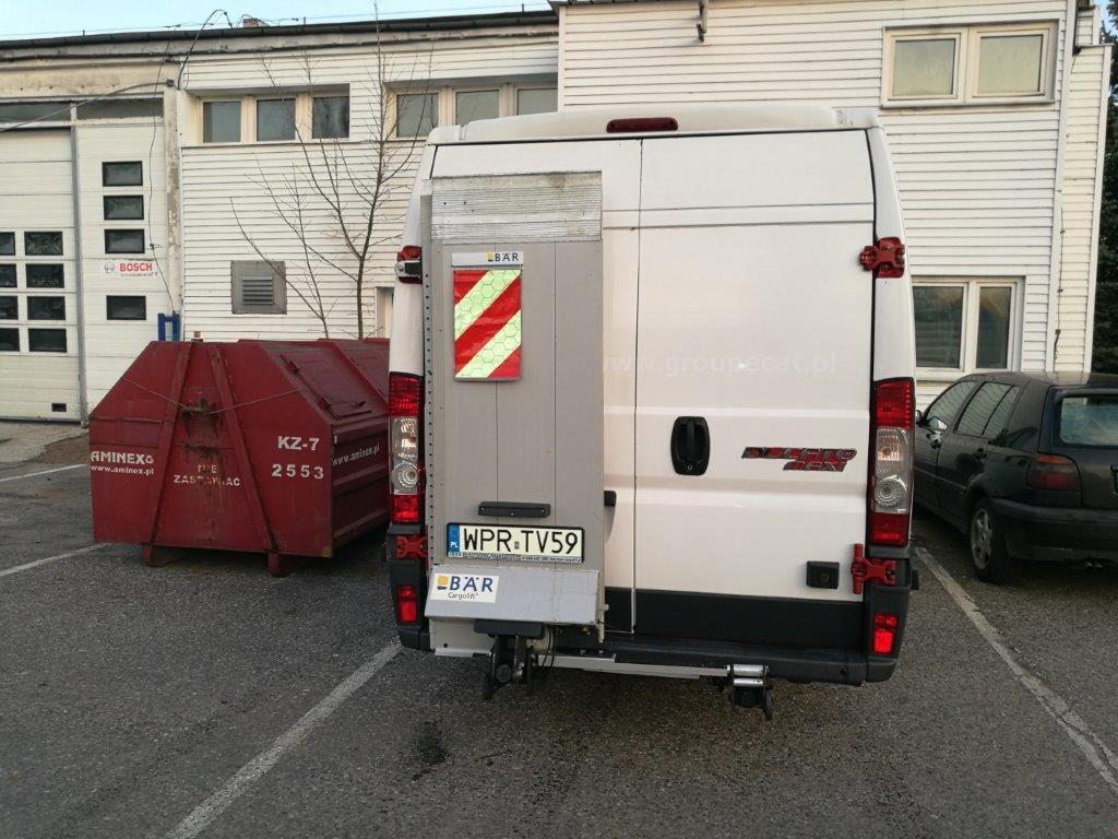 Tył białego busa dostawczego z otwartymi drzwiami i rampą, zaparkowanego na parkingu przed budynkiem przemysłowym, obok stoi czerwony kontener na śmieci.