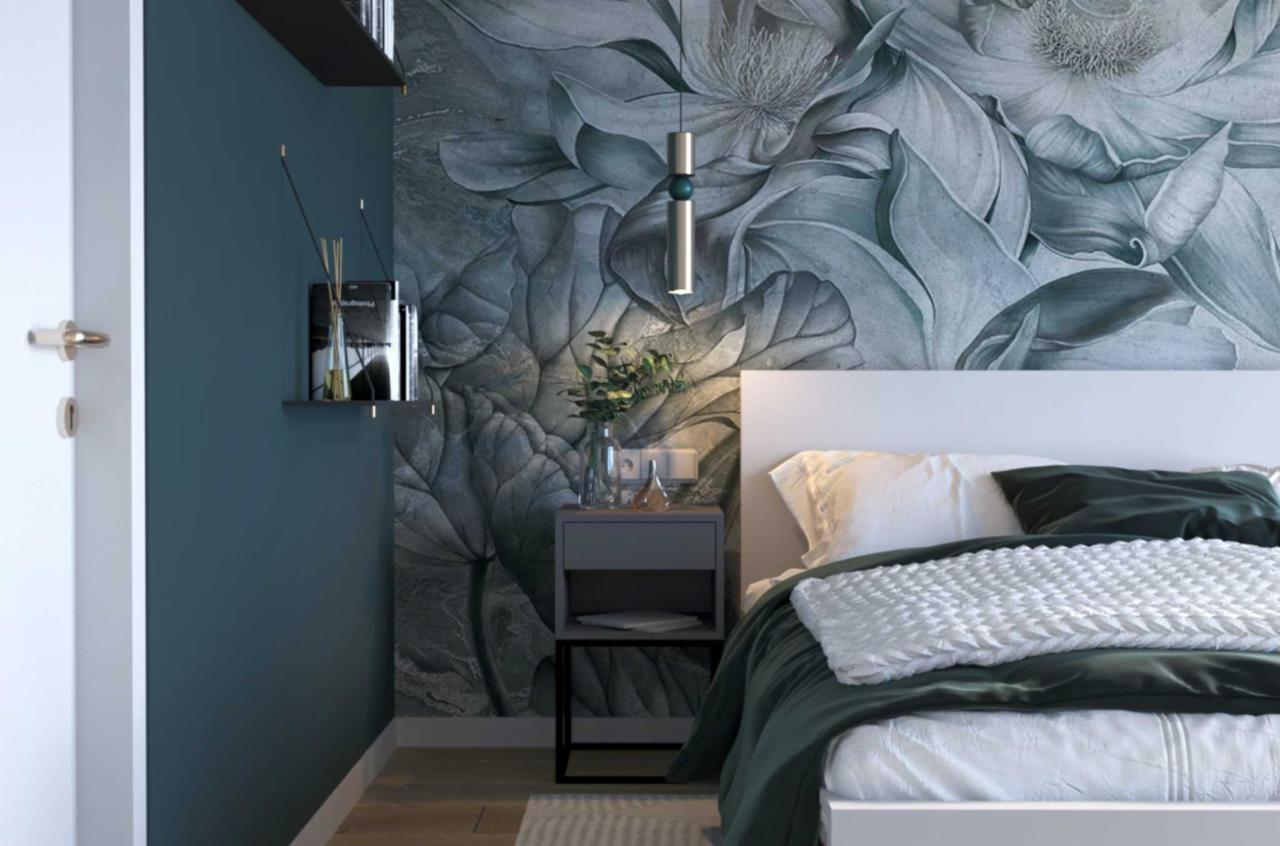 Sypialnia z tapetą w motywy roślinne, białe łóżko z zieloną narzutą, szara szafka nocna z lampką i wazonem, wisząca lampa z zielonym akcentem, półki na ścianie.