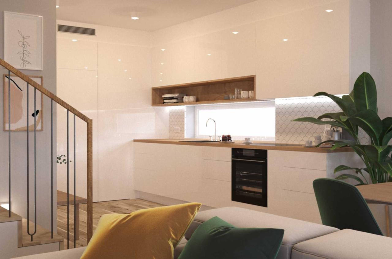 Jasna kuchnia z białymi szafkami, drewnianym blatem i otwartą półką, widoczna z perspektywy salonu z schodami i rośliną doniczkową.