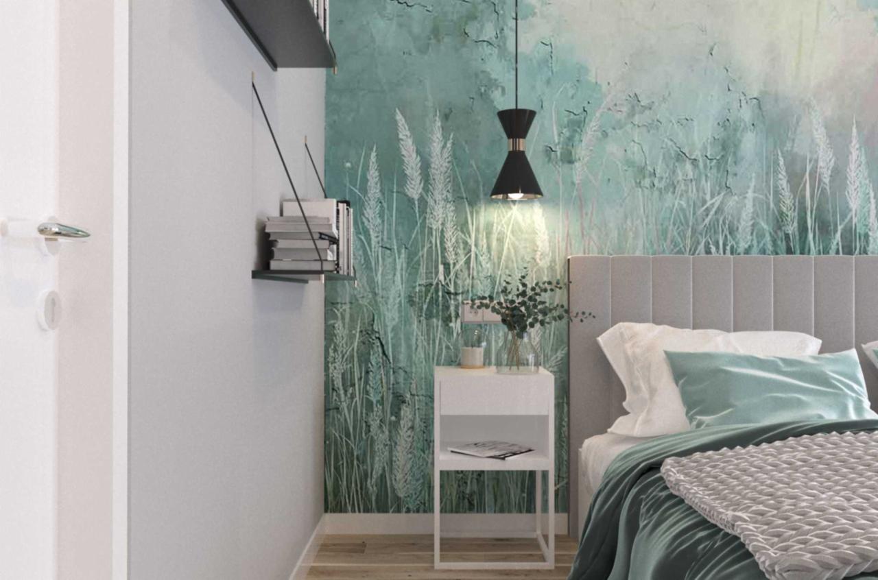 Sypialnia z łóżkiem z szarym wezgłowiem, białą szafką nocną z dekoracjami, lampą wiszącą i tapetą w motywie łąki w odcieniach zieleni i bieli.