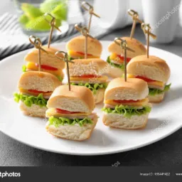 Mini kanapki z sałatą, pomidorem i serem na białym talerzu, spięte ozdobnymi wykałaczkami.