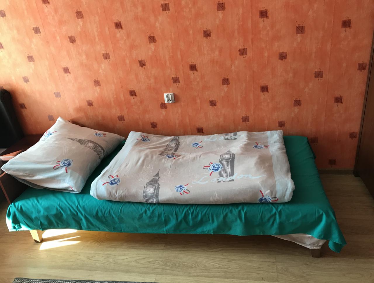 Rozłożona sofa z zielonym obrusem i pościelą z motywem Big Bena i róż na pomarańczowej ścianie z małymi kwadratami.