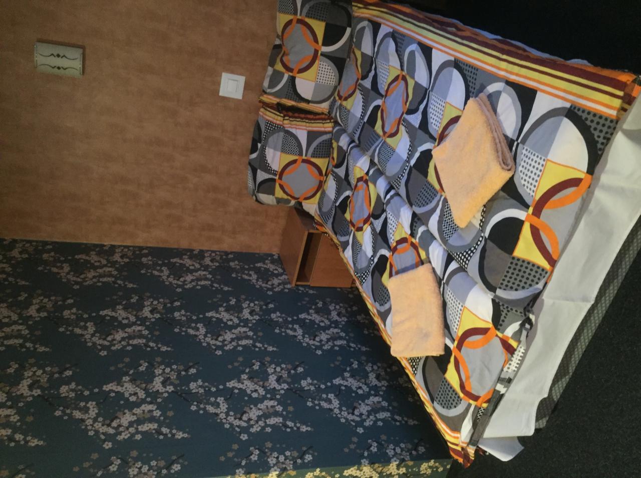 Pokój hotelowy z łóżkiem w geometryczne wzory i dwoma ręcznikami, brązowa ściana oraz dywan w drobne kwiatki