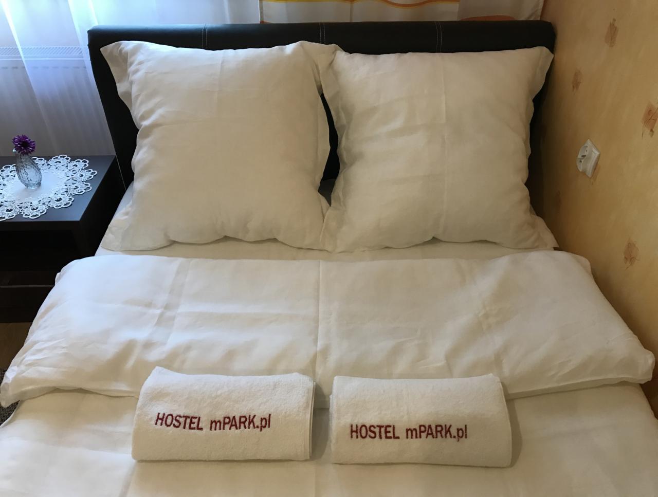 Pokój z dwoma poduszkami i ręcznikami z napisem 'HOSTEL mPARK.pl' na łóżku z czarnym zagłówkiem, stolik nocny z kwiatami po lewej stronie.