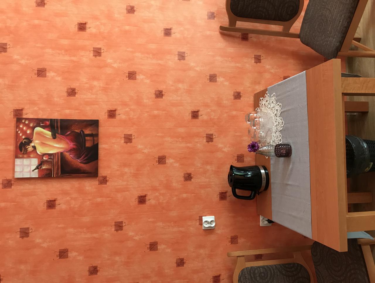 Pomarańczowa ściana z wzorem, na której wisi obraz z kobiecą sylwetką. Obok stół z nakryciem, na którym stoją szklanki, dzbanek i dekoracja kwiatowa. Widoczne krzesła i gniazdko elektryczne.