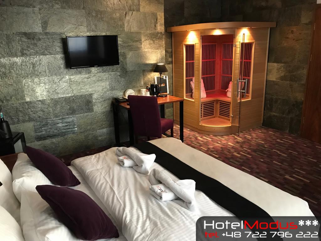 Pokój hotelowy z prywatną sauną infrared, łóżko z purpurowymi poduszkami i ręcznikami, ściana z kamienia naturalnego, telewizor LCD i biurko z czajnikiem.