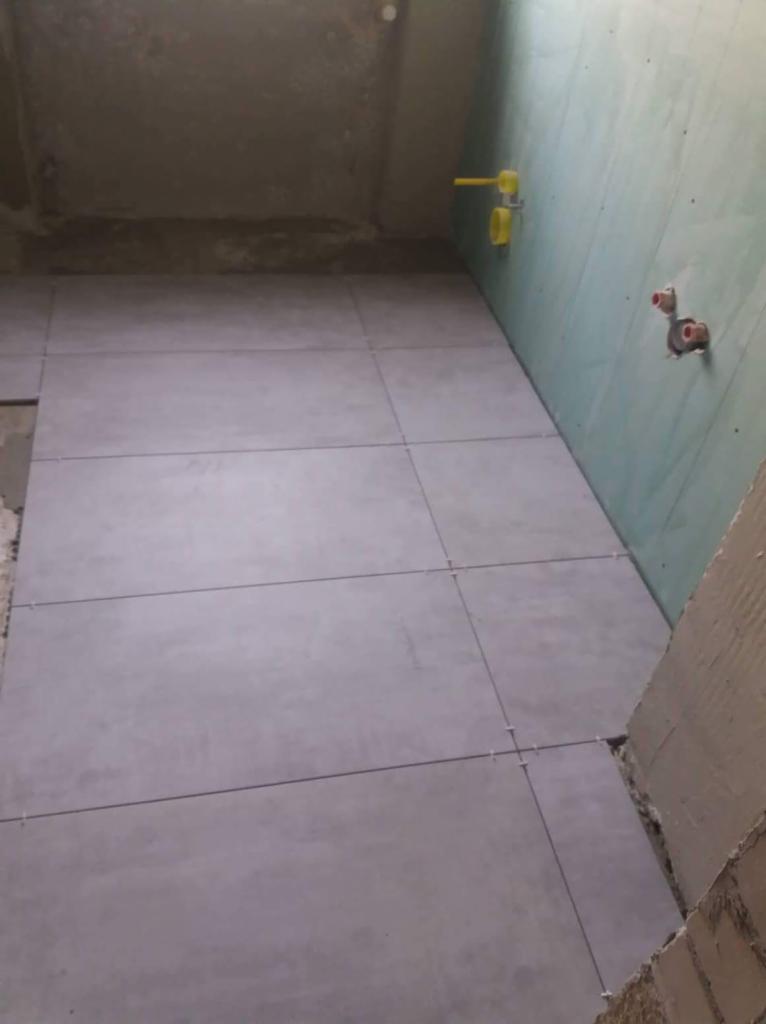 Szare, matowe płytki podłogowe ułożone w łazience, widoczne krzyżyki dystansowe, ściany przygotowane pod dalsze prace wykończeniowe, instalacja wodna wystaje ze ściany.