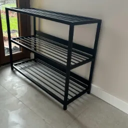 Trzypoziomowy, czarny, metalowy regał na buty, widoczny na tle jasnej ściany i fragmentu okna z zielenią.