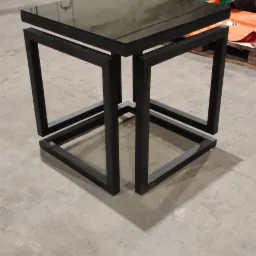 Stolik kawowy z czarnym szklanym blatem i geometryczną podstawą z profili stalowych, wykonany w stylu industrialnym, na tle betonowej posadzki w warsztacie.