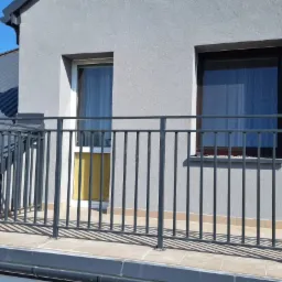 Szara, metalowa barierka balkonowa z pionowymi szczeblami na tle szarej ściany budynku z dwoma oknami i fragmentu dachu pokrytego blachą trapezową.