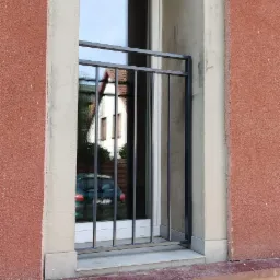 Czarna, metalowa balustrada okienna z pionowymi prętami, zamontowana w oknie w trakcie wykańczania elewacji budynku o czerwonej, chropowatej strukturze, z widocznym odbiciem samochodu i domu w szybie.