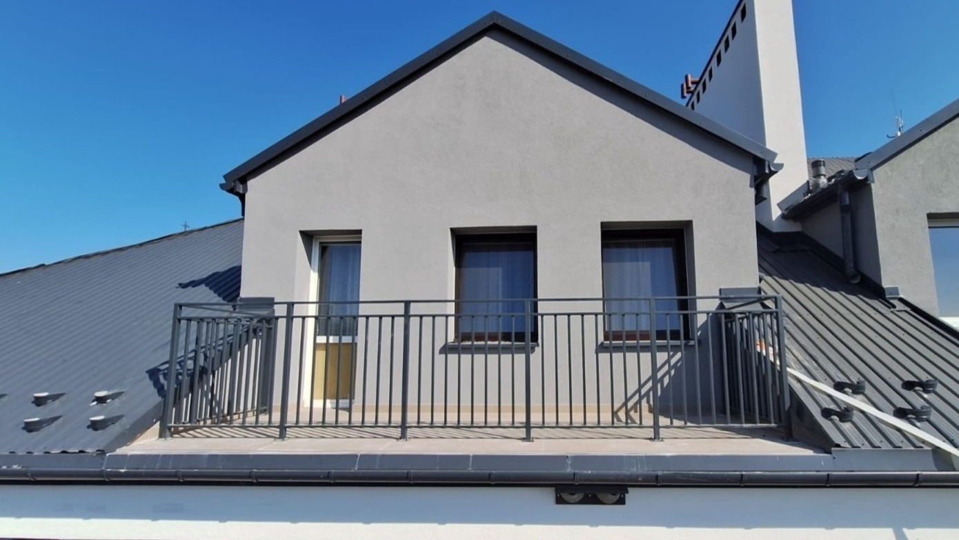 Balkon z czarną, metalową balustradą na dachu budynku z szarą elewacją i trzema oknami pod skośnym dachem pokrytym blachą.