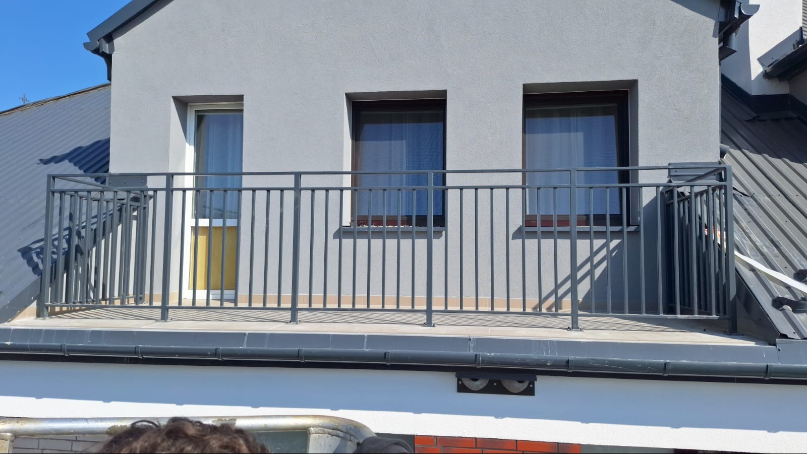 Szara, metalowa balustrada balkonowa z pionowymi szczebelkami, zamontowana na tarasie na dachu budynku z trzema oknami o różnych rozmiarach.