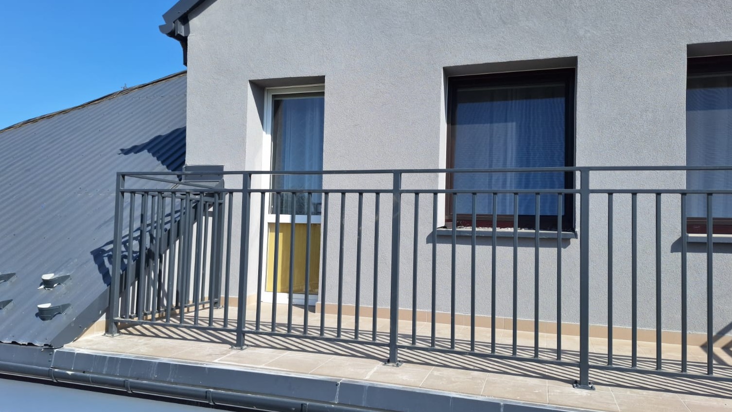 Szara, metalowa barierka balkonowa z pionowymi szczeblami na tle szarej ściany budynku z dwoma oknami i fragmentu dachu pokrytego blachą trapezową.