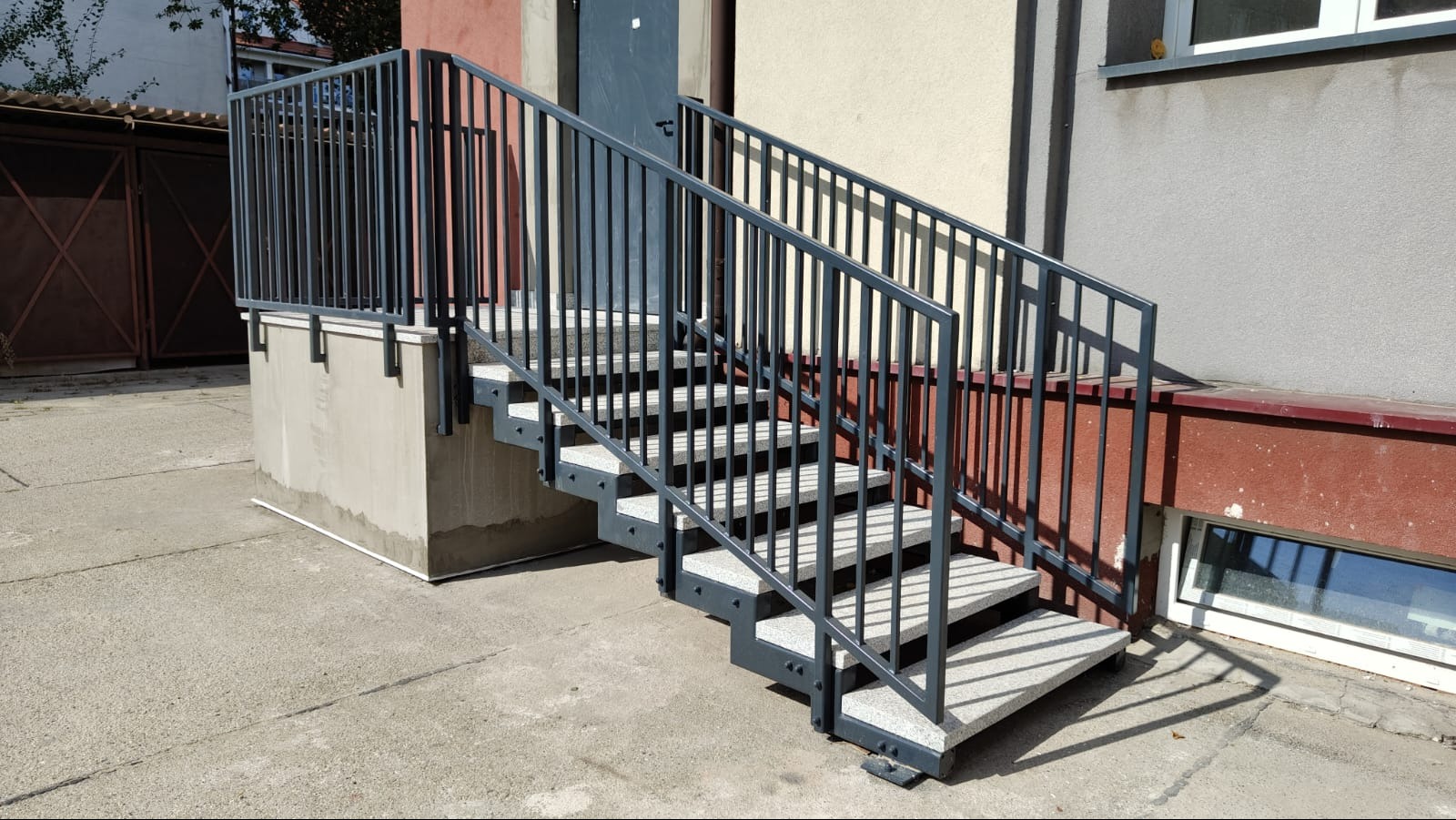 Zewnętrzne schody z szarymi, metalowymi poręczami i balustradą, prowadzące do wejścia budynku.