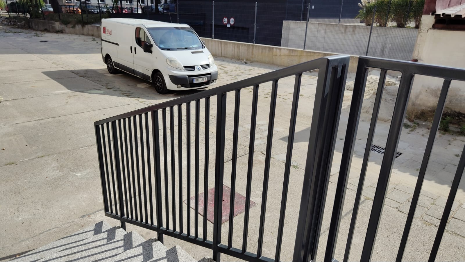 Czarne metalowe barierki schodowe z widocznym samochodem dostawczym firmy w tle na betonowym placu.