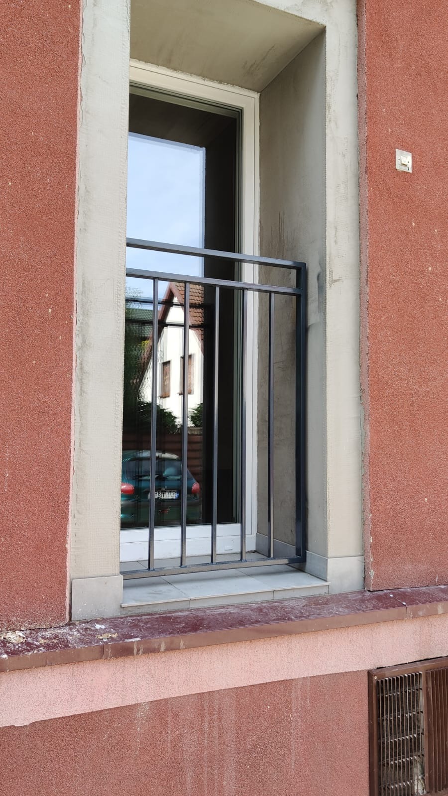 Czarna, metalowa balustrada okienna z pionowymi prętami, zamontowana w oknie w trakcie wykańczania elewacji budynku o czerwonej, chropowatej strukturze, z widocznym odbiciem samochodu i domu w szybie.