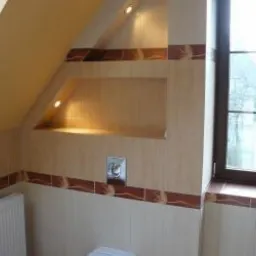 Ukończona łazienka na poddaszu z beżowymi płytkami, dekoracyjnym brązowym pasem i wnęką z oświetleniem halogenowym nad spłuczką. Widoczne okno i grzejnik.