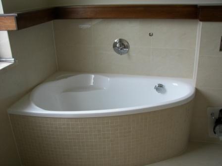 Narożna wanna obudowana mozaiką w beżowych odcieniach, zamontowana bateria ścienna, kremowe płytki na ścianach, drewniana listwa podsufitowa.