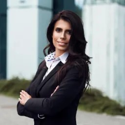 Uśmiechnięta kobieta w czarnym żakiecie i białej koszuli z wzorem kotwic, stojąca na tle nowoczesnego biurowca z zielenią.