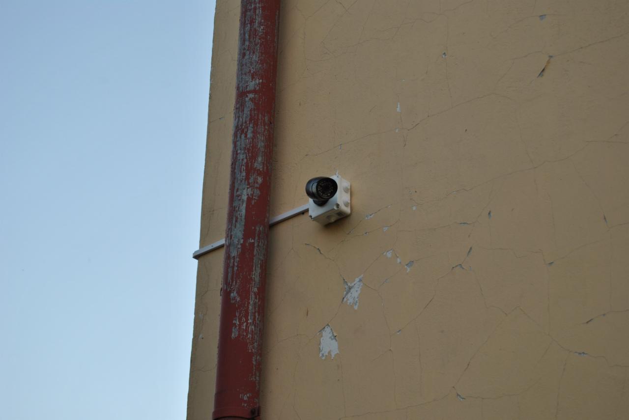 Zewnętrzna kamera monitoringu przymocowana do ściany budynku, obok rury spustowej z odpryskującą farbą.