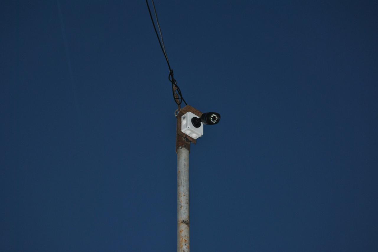 Kamera monitoringu na metalowym słupie z białą obudową, widoczna na tle intensywnego, błękitnego nieba.