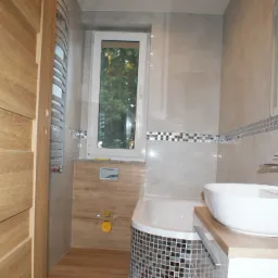 Nowoczesna łazienka z beżowymi płytkami, mozaiką i drewnianymi elementami, owalną umywalką na blacie i częściowo obudowaną wanną z mozaiką, widok na okno z zielenią.