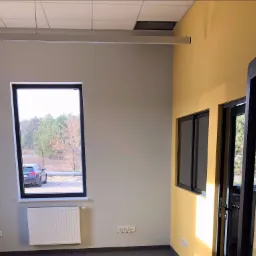 Świeżo pomalowane wnętrze biura z szarą i żółtą ścianą, czarna rama okienna z widokiem na parking i samochód, biały grzejnik pod oknem, szara wykładzina dywanowa.