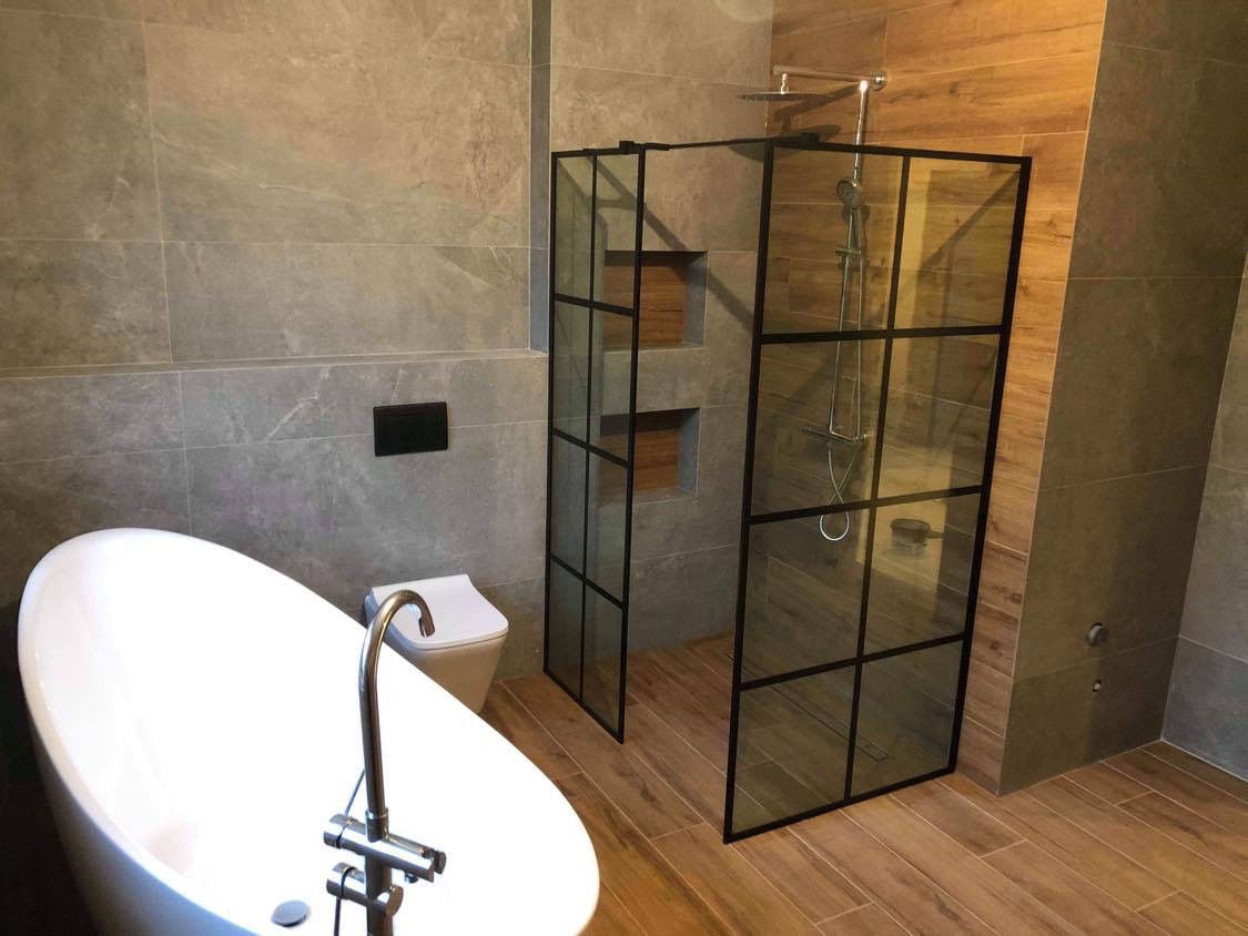 Nowoczesna łazienka z wolnostojącą wanną, toaletą i kabiną prysznicową w stylu industrialnym z czarnymi ramami, wykończona płytkami imitującymi kamień i drewno.