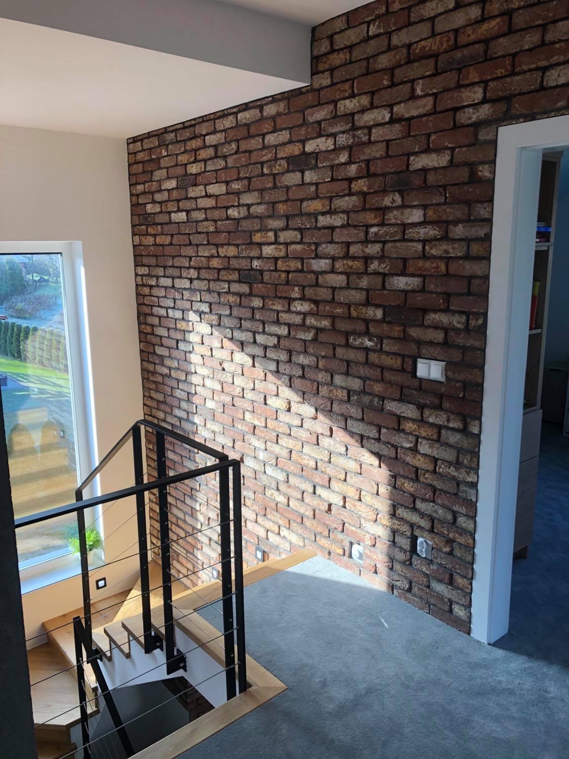 Ściana z cegły dekoracyjnej w korytarzu z widocznymi schodami i oknem, oświetlona naturalnym światłem tworzącym kontrast cieni.