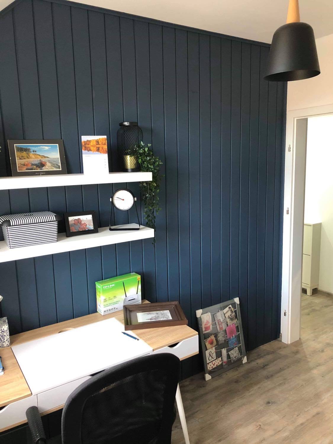 Ściana z ciemnoniebieskich paneli pionowych, białe półki z dekoracjami, biurko z akcesoriami, drewniana podłoga, lampa wisząca, widok na inne pomieszczenie przez otwarte drzwi.