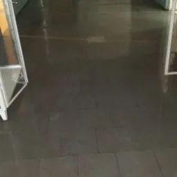 Mokre, świeżo umyte płytki podłogowe w korytarzu szpitalnym, widoczny fragment wiadra z płynem i żółta szmatka.