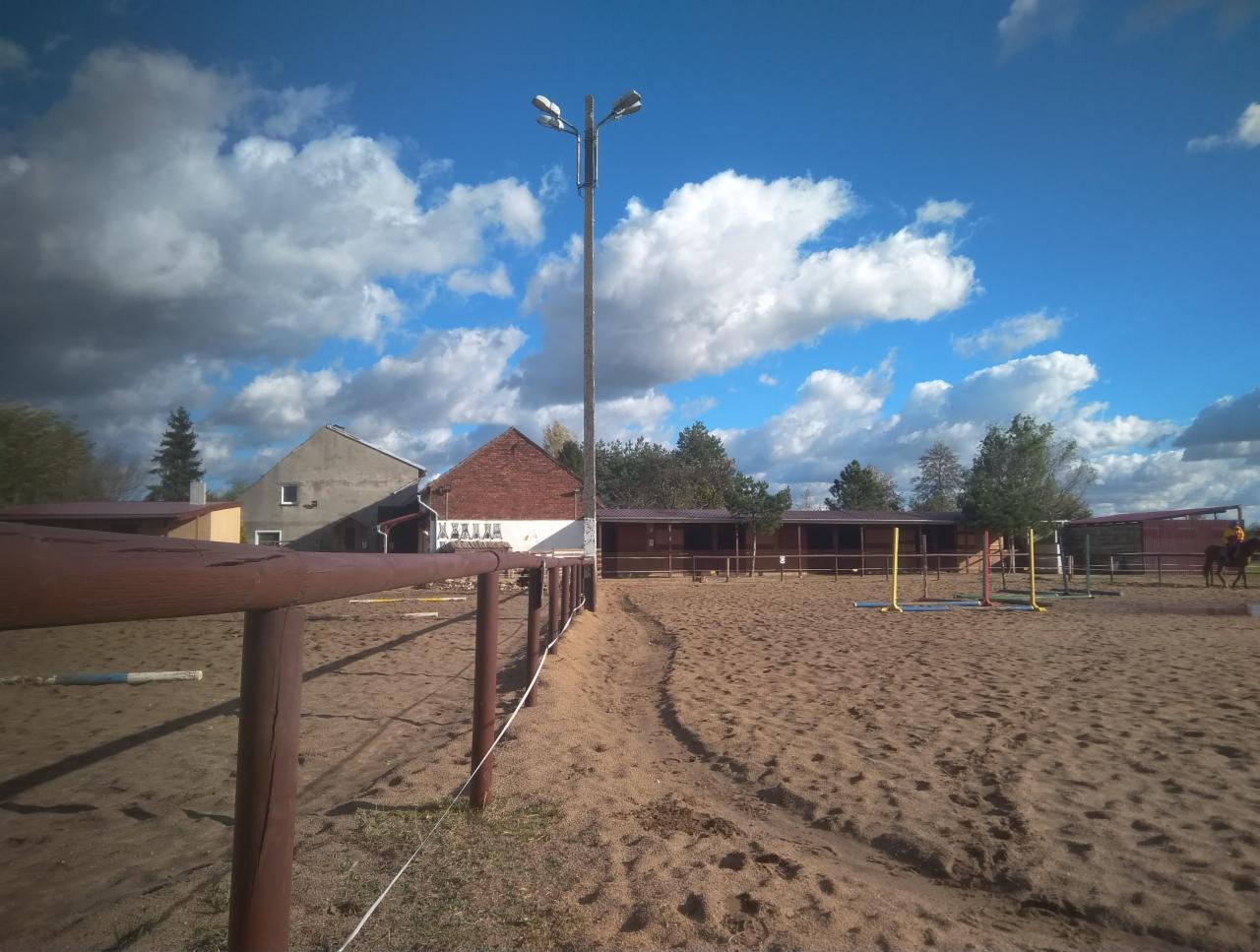Piaszczysty plac do jazdy konnej z przeszkodami, widok na budynki gospodarcze i błękitne niebo z chmurami.