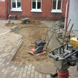 Układanie kostki brukowej na terenie posesji z ceglanym budynkiem w tle, widoczne ubijarki i przygotowany teren z piaskiem.