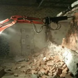Pomarańczowa minikoparka Kubota z hydraulicznym młotem pneumatycznym rozbija ceglany mur wewnątrz budynku, gruz leży na podłodze, widoczne ślady po instalacjach na ścianie w tle.
