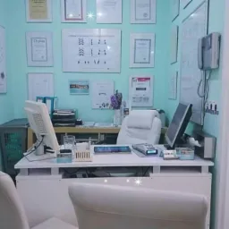 Gabinet lekarski z biurkiem, dwoma monitorami, krzesłem z białej skóry i ścianami ozdobionymi dyplomami i plakatem anatomicznym.