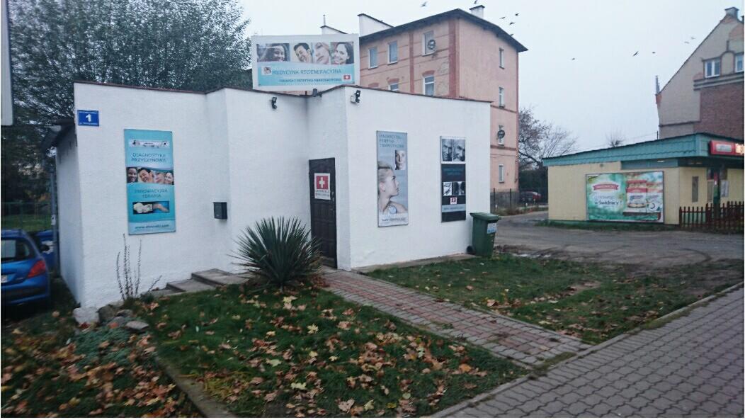 Biały budynek kliniki z reklamami na ścianach, trawnikiem z opadłymi liśćmi i zielonym koszem na śmieci przed wejściem, widziany z perspektywy ulicy w pochmurny dzień.