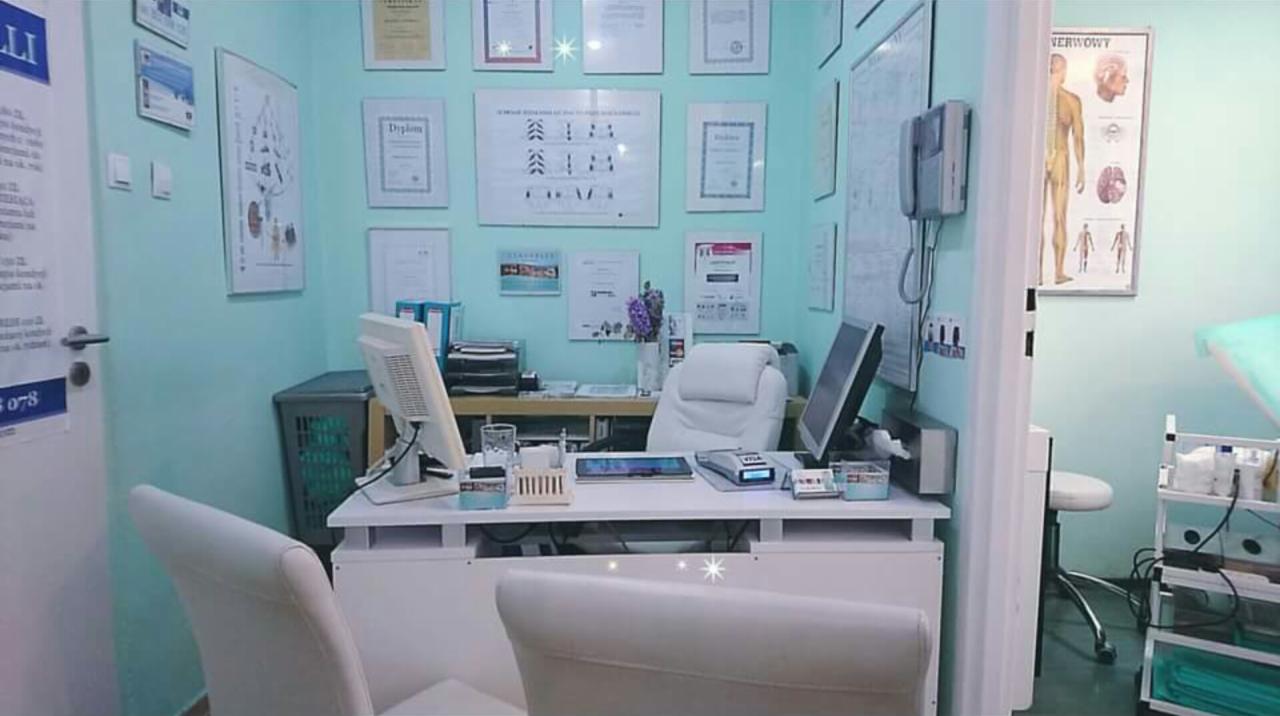 Gabinet lekarski z biurkiem, dwoma monitorami, krzesłem z białej skóry i ścianami ozdobionymi dyplomami i plakatem anatomicznym.
