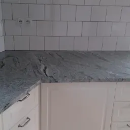 Szary blat kuchenny z wyraźnym wzorem kamienia, zamontowany na białych szafkach z chromowanymi uchwytami, tło stanowią białe kafelki ułożone w cegiełkę.